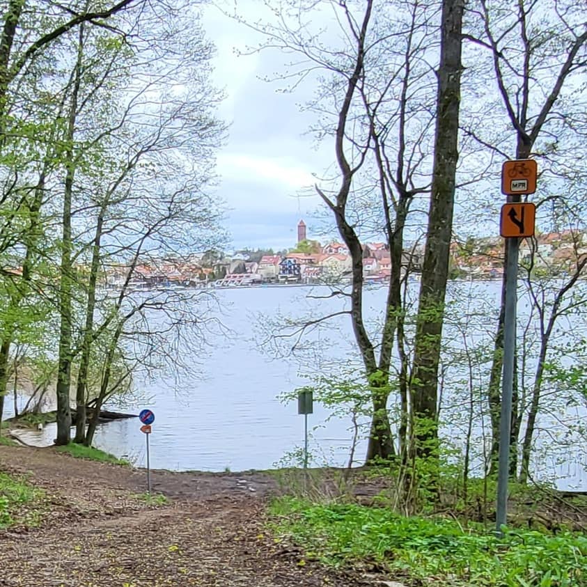 Widok na Mikołajki z trasy Mazury – Pętla Rowerowa, z oznaczeniami szlaku rowerowego wśród drzew nad jeziorem.