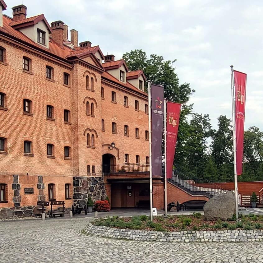 Zamek w Rynie na Mazurach, historyczna budowla otoczona brukowanym placem – atrakcja w regionie Mazurskiej Pętli Rowerowej.